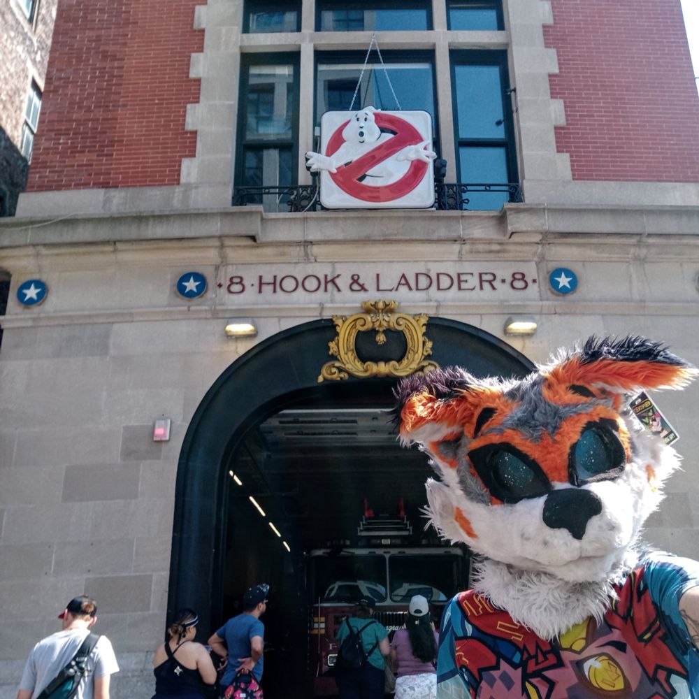 An orange, silver, and white fox fursuiter in front of the Ghostbusters firehouse 