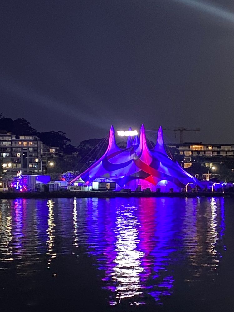 A brightly lit blue and red circus tent reflects in the dark evening ripples of Gosford waterfront