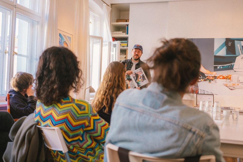 Nikita Teryoshin, wearing a blue cap, standing in front of a small audience in a bright room, holding an open book. The listeners, seated on chairs, face him attentively. A large photograph hangs on the wall behind him.