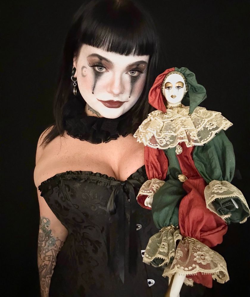 creepy is in clown cosplay wearing a black corset and holding up her haunted porcelain clown doll. creepy did her makeup to match the doll's. 
