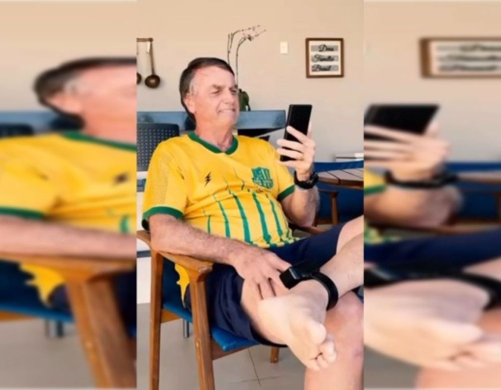 O ex-presidente e agora presidiário, Bolsonaro, sentado com a camisa do Brasil e uma tornozeleira eletrônica. 