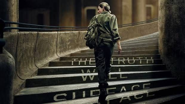 A woman with her back to the viewer ascends a wide, stone staircase. She is dressed in a dark green, utilitarian jacket and trousers, with a backpack slung over one shoulder. The lighting from above highlights the worn texture of the concrete steps and walls. White, block letters are painted onto the steps, partially obscured by her movement, but clearly spell out "THE TRUTH WILL SURFACE". The overall atmosphere is somber and somewhat mysterious.