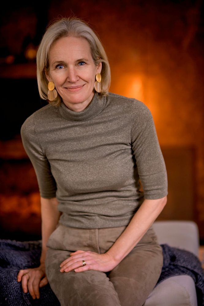 Picture of Cokky Hilhorst. Cokky is the new dean of the faculty of Technology, Policy and Management. In this picture, she is sitting on a grey ottoman, with one of her hands resting on her leg. Coccky is wearing a beige-grey turtleneck top and brown trousers. She is looking into the camera and has a smile on her face.