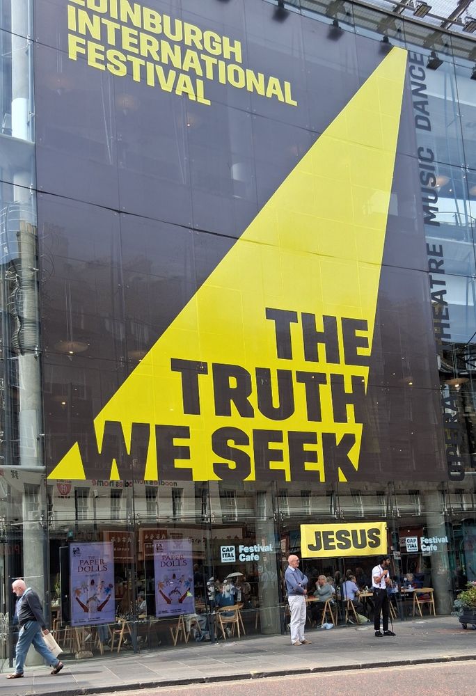 The Edinburgh International Festival branding on the side of the Festival Theatre, which says THE TRUTH WE SEEK in big black on yellow letters. Underneath it, a couple of lads hold up a banner in the same style which says JESUS