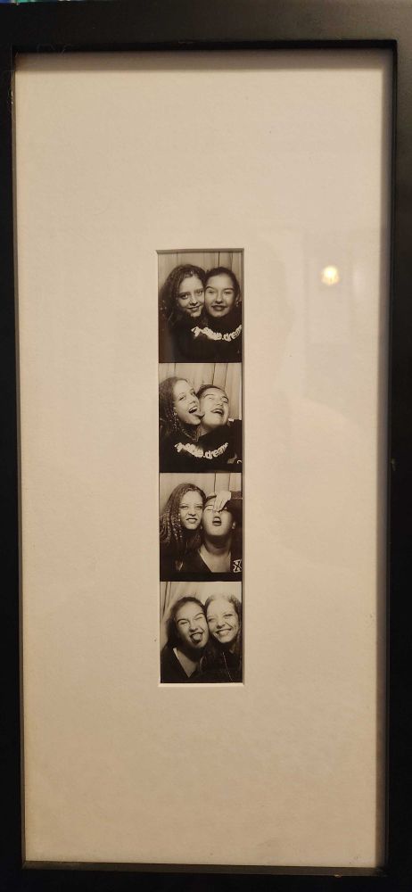 A framed black and white photobooth strip of two young women, one with long blondeish hair, and one with dark hair tied up in a bun. They make silly faces and hug each other.