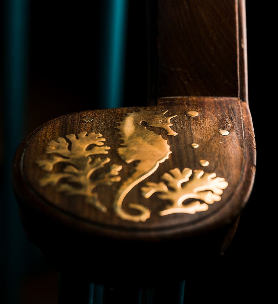 a colour photograph of the brass inlay of a staircase finial. The inlay shows a seahorse, surrounded by two fronds of seaweed and some bubbles 