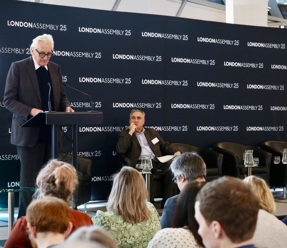 Photo of man speaking at podium with a man behind sat down and the backs of a crowd's heads