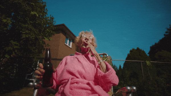 an elderly woman in a pink robe outside in a patio chair with a beer and a blunt