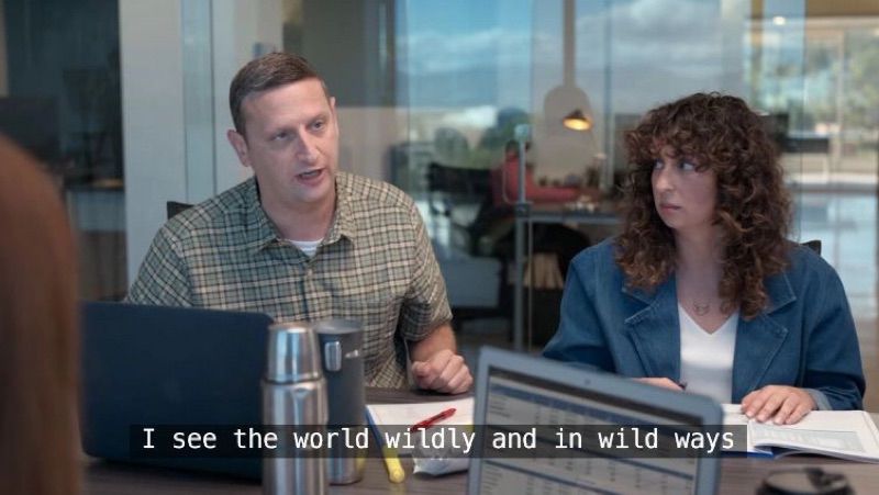 Tim Robinson sitting at a conference table with a laptop in front of him, saying “I see the world wildly and in wild ways”. A woman sits next to him and is looking at him.
