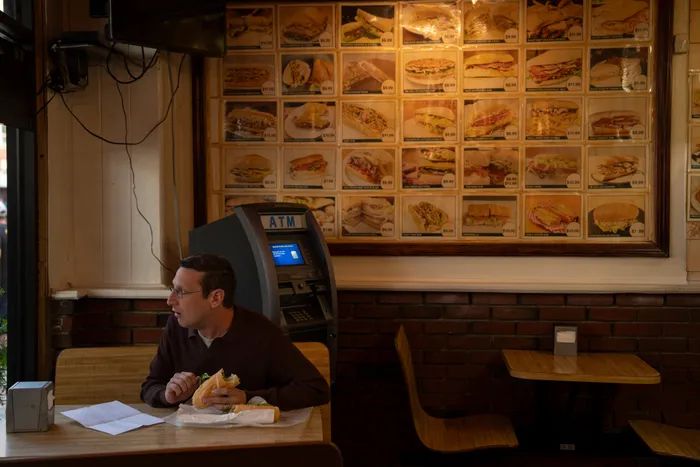 Tim Robinson sitting in the booth of a sandwich restaurant alone, eating a sandwich and staring out the window.