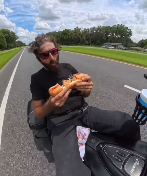 A man on a motorcycle eating a meatball sub - by taking bites out of it sideways instead of starting at one end and working his way down, like a civilized person.