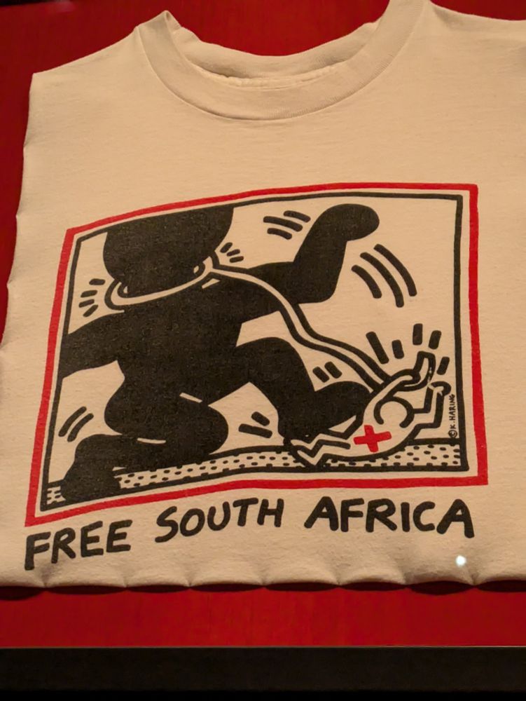 black red and white Keith Haring image created against South Africa apartheid. a large bond person has a rope around their neck held by a small white person and the black person is stepping on the white person.