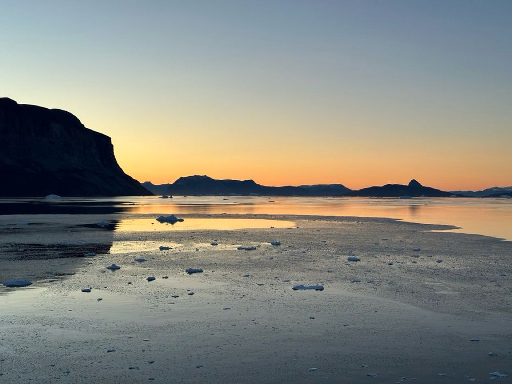 Ein stiller Sonnenuntergang über einer gefrorenen Meereslandschaft mit verstreuten Eisschollen, dunklen Felsen und sanft schimmerndem Himmel in warmen Orangetönen.