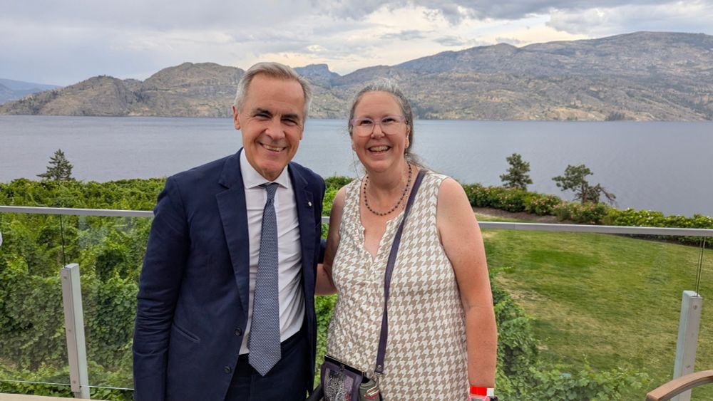 An older man with a dark blue suit and a grey tie stands beside a middle-aged woman with long hair in a ponytail and light pink glasses wearing a checked beige and white vest. Behind them is a vineyard, and large lake and some low mountains.