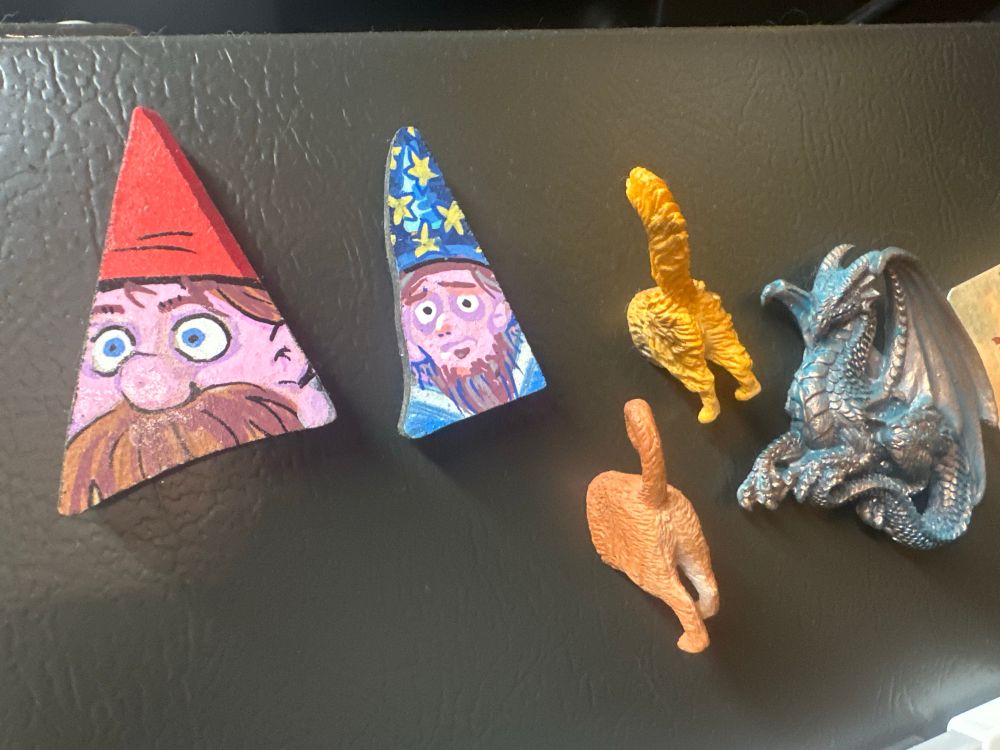 Photo of handmade gnome magnets on a refrigerator. One has a red hat, and the other has a blue hat with stars. They’re placed beside two cat butt magnets and one of a dragon