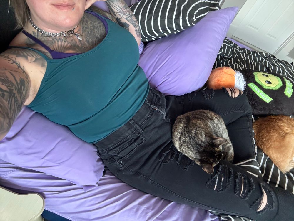 Strata the torbie cat curled up between Riley’s legs, Hobbes the orange and white cat curled up nearby, Riley in a teal cami and distressed black jeans 