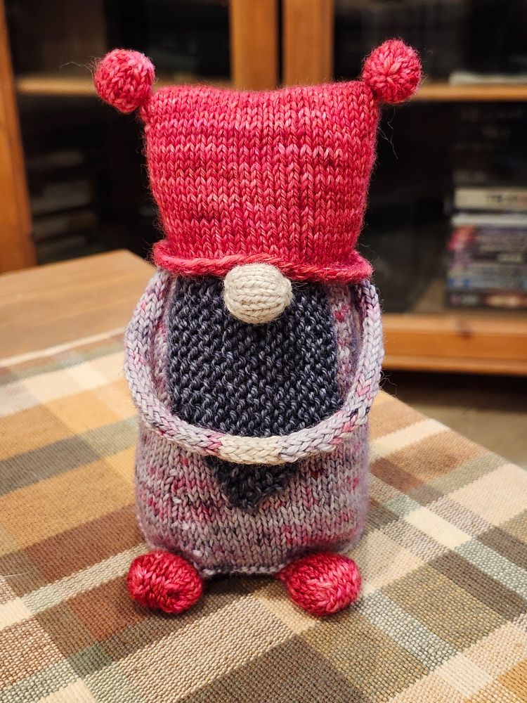 A handknit gnome, clasping his hands in front of his beard. Pink hat & shoes, speckled pink & grey body, dark grey beard.