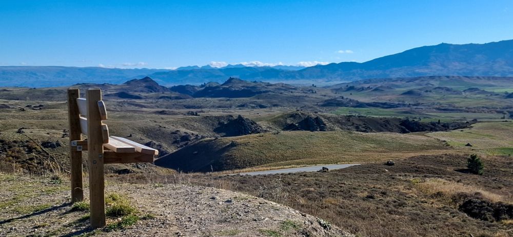 Bench overlooking Manuherikia valley with Dunsta and Old Man range with Remarkeble Mountains in distance, under blue sky.