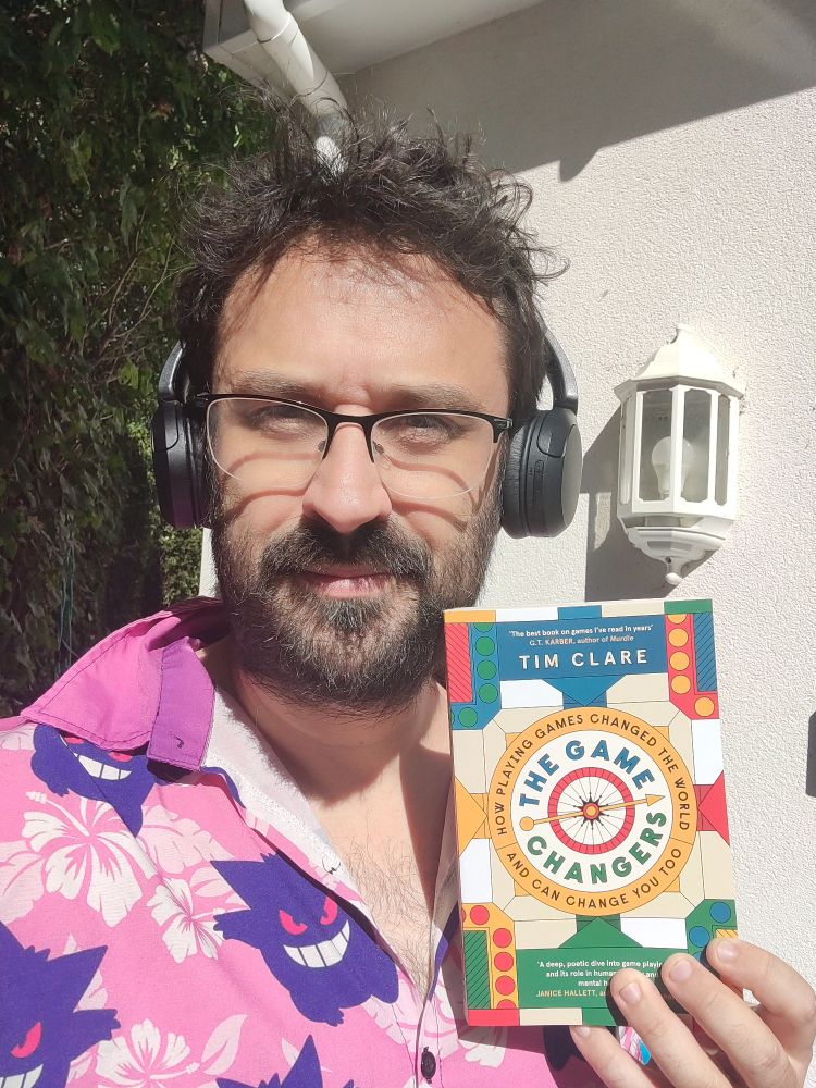 A haggard hirsute bespectacled Tim Clare (me) in a pink Gengar shirt holding my book, The Game Changers. I look less old when I've had a shave