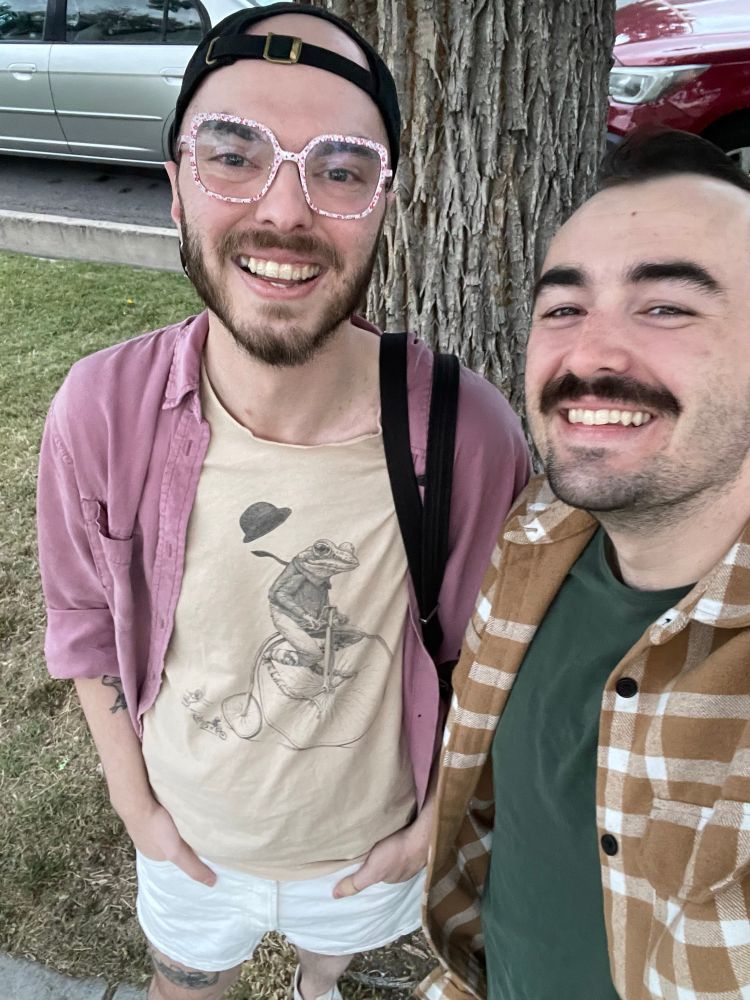 Benny boi is dressed in a tan shirt with a frog riding a high wheel bicycle with its hat flying off. Layered on top of their t-shirt is a plum colored button up. Benny has a goofy grin on their face because we had just run into the tree behind us. To the right of Benny is their boyfriend in a green tee with a yellow flannel layered on top. They also have a goofy grin on their face, mostly because Benny’s fit is exactly why we both got clocked as being fruity by the shoe store employee.