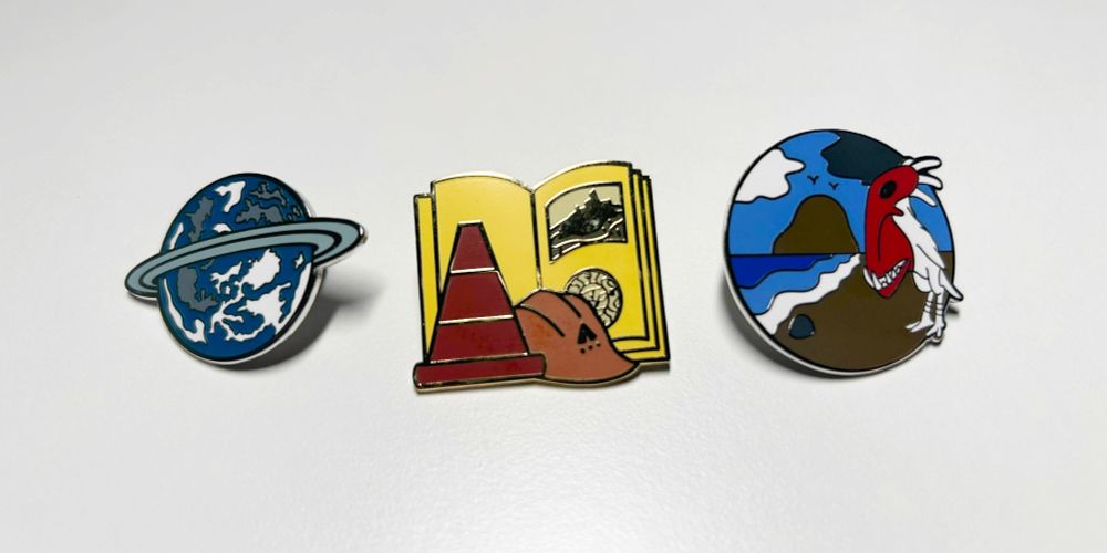 Myst-themed enamel pins depicting Todelmer, a linking book with a cone and D'ni Restoration Council hard hat, and a piranha chicken on a beach.