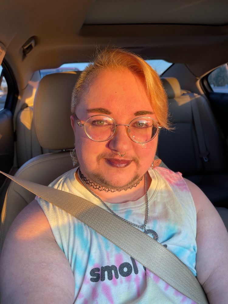 A blond trans man. He has a dark goatee. He’s sporting round golden glasses and a gold septum ring. His hair is combed to the side, and he’s wearing a tie-dye top that reads “Smol Gay”. He’s also wearing a black plastic choker and a silver necklace that reads “my sun and stars”. His earrings are death’s head moths and he’s got on purple lipstick and eye shadow. His brows are also filled out. 