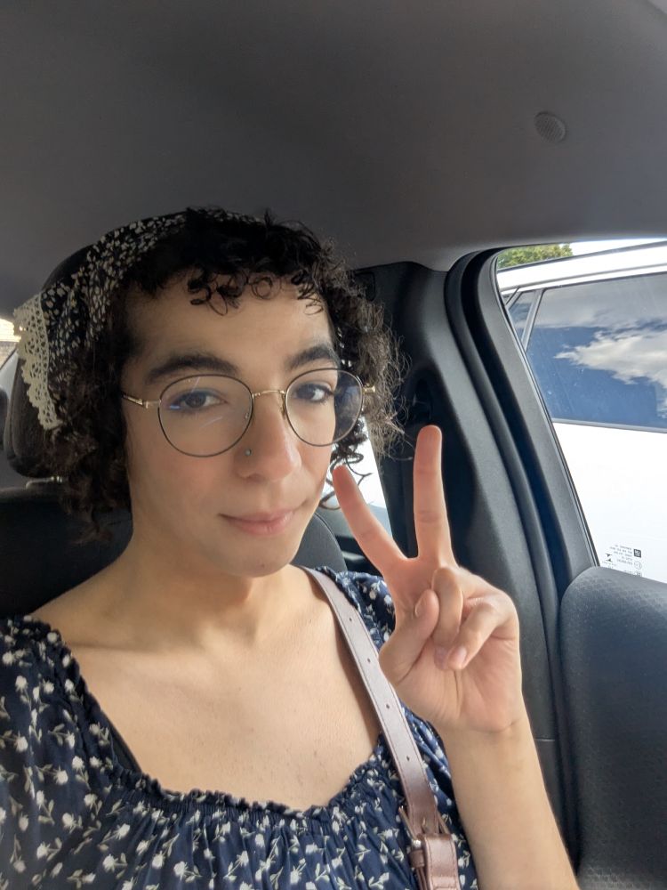 Car selfie of me wearing a dark blue dress with white flowers and a blue and white bandana, giving peace sign