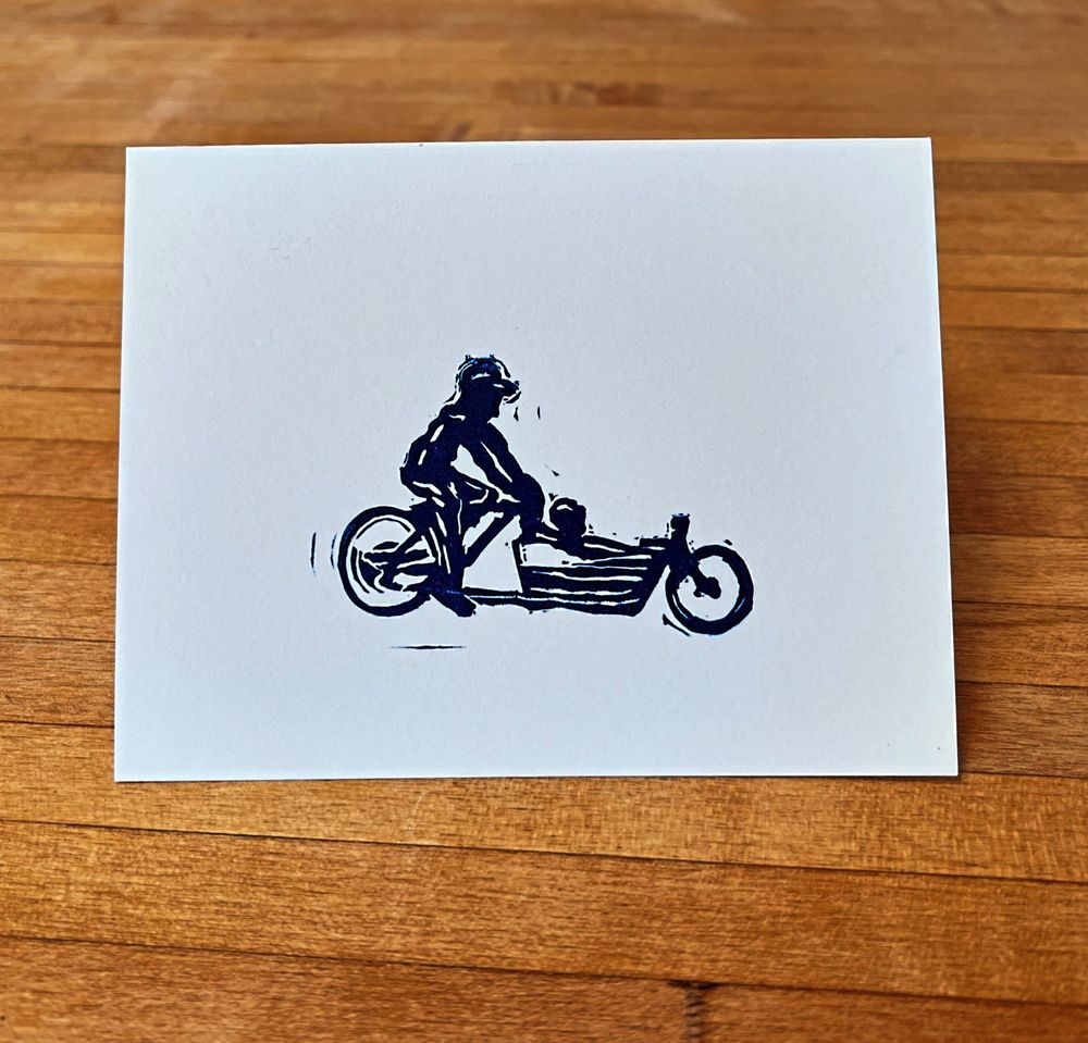 Card with a blue ink print of a person ridding a bakfiets/ cargo bike with a kiddo in the front. 
