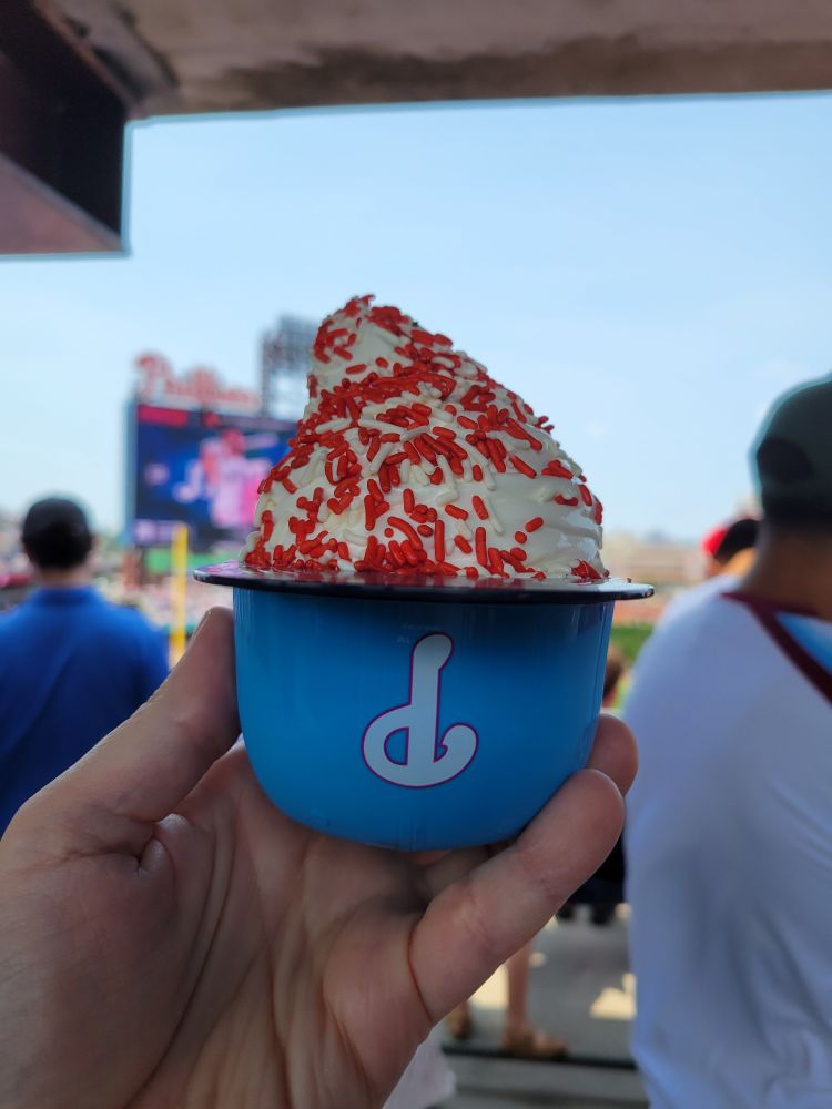 It's soft vanilla ice cream in a plastic miniature batting helmet. Red and white sprinkles top the ice cream. The helmet is light blue, with a dark blue brim. The crown bears a stylized letter P, in white.