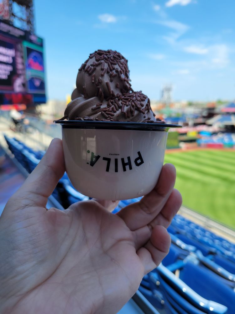 It's soft chocolate ice cream in a plastic batting helmet. The ice cream is topped with chocolate sprinkles. The helmet is cream-colored, with a black brim. "Philadelphia" is abbreviated, in black, on the crown.