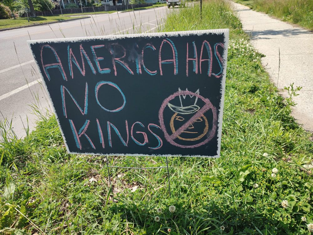 A gray chalkboard sign that reads "AMERICA HAS NO KINGS" in blue and red lettering. Inside a big red circle with a red line through it is an orange face with empty eyes and a recognizable toupe of blond hair and a crown. 