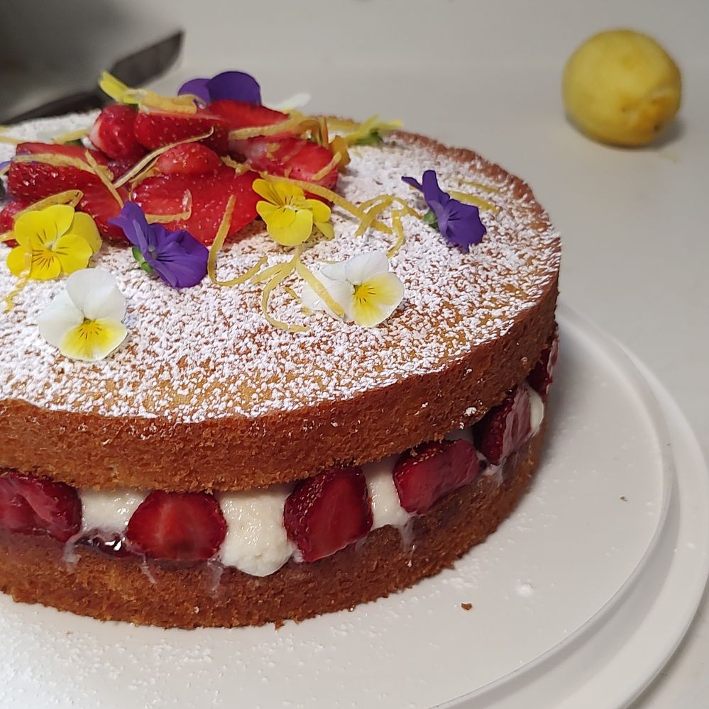 Two layers of vanilla cake with strawberries and cream layered visibly in between. The top is sprinkled with powdered sugar and scattered on top are sliced strawberries, violets in purple yellow and white, and bits of lemon zest. The cake is photographed from the side and slightly above so you can see both the filling and topping. 