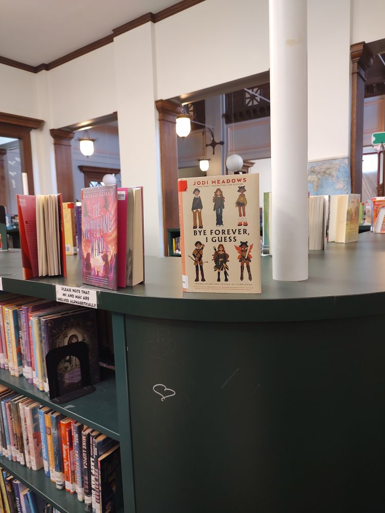 A green library shelf with new books displayed on top cover facing out. Front and center is "Bye Forever, I Guess" by Jodi Meadows. 