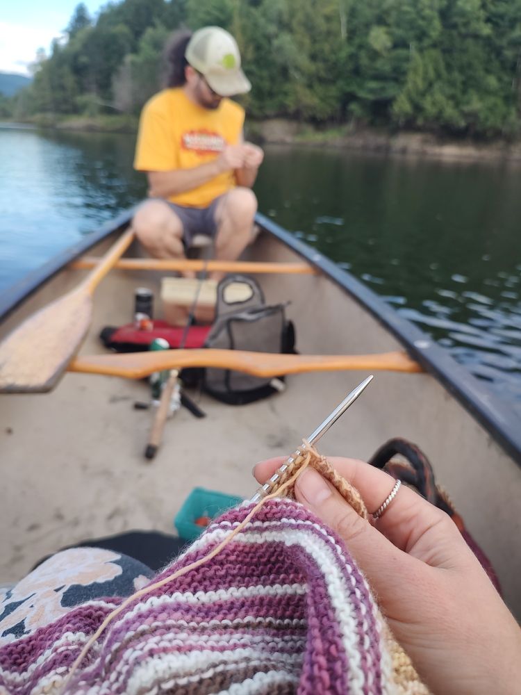 The point of view is from a canoe seat, looking across the boat at the other paddler. Foreground shows a pink, white, and gold garter stitch work in progress with one hand holding up the needles. In the out-of-focus background a man in a hat fiddles with his fishing pole. 