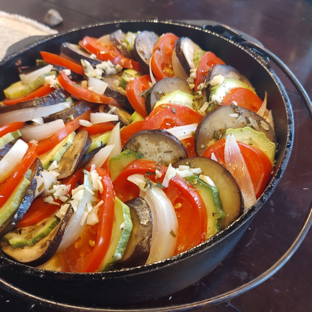 A cast iron dutch oven full to the brim. Slices of zucchini, eggplant, tomato, and onion are layered in a spiral on the top and sprinkled with minced garlic and herbs. 