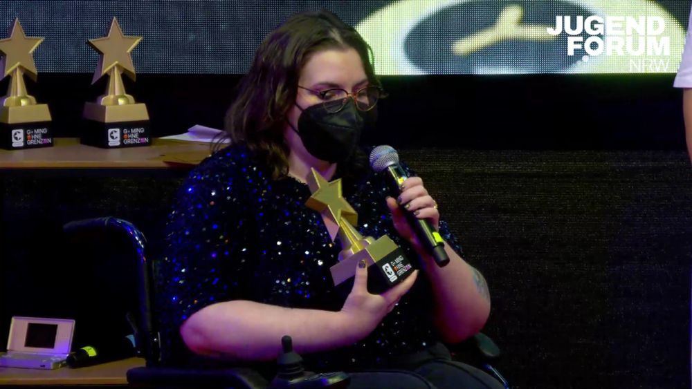 Radderss, a disabled woman, on stage holding a large golden star-shaped award, and speaking into a microphone. 
