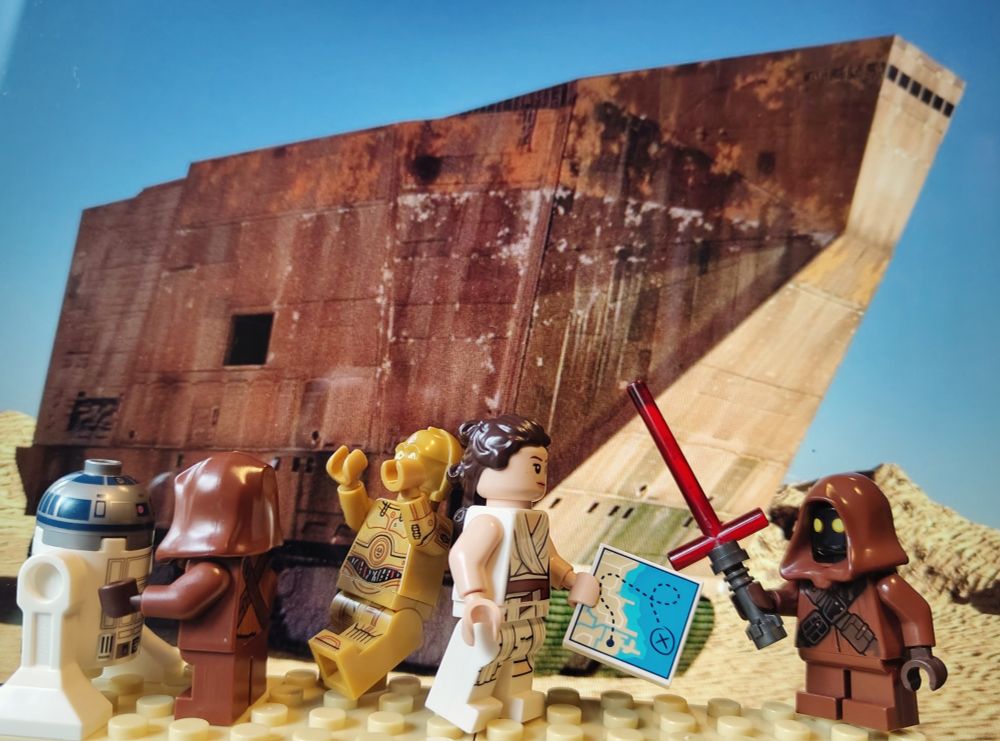 Lego figures of R2D2, C3PO, Rey and Jawa's in front of a sand crawler on Tatooine. On Jawa has Kylo Ren's light saber. 