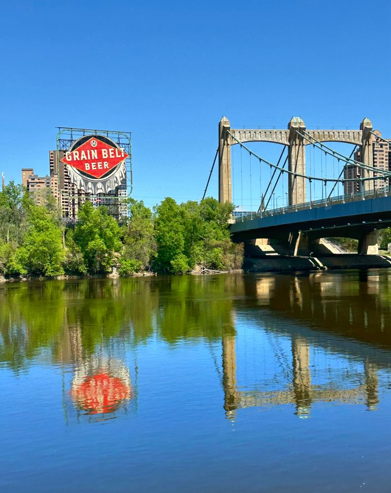 Calm waters and a sunny day make for a reflection of the Grain Belt sign and the Hennepin Ave bridge on the Mississippi River.