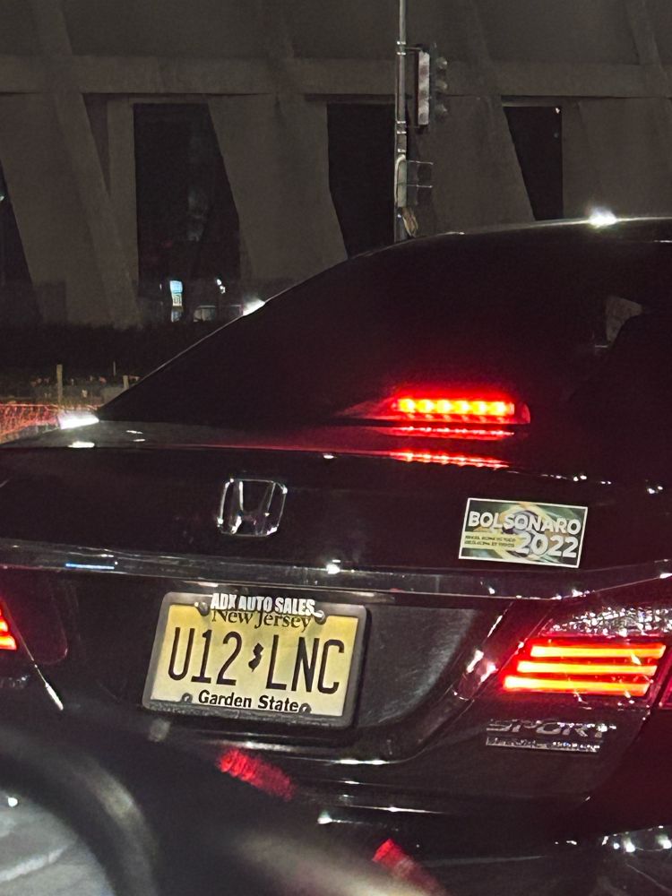 A zoomed in photo of the back of a black car. The car has a license plate from New Jersey and a Bolsonaro 2022 bumper sticker