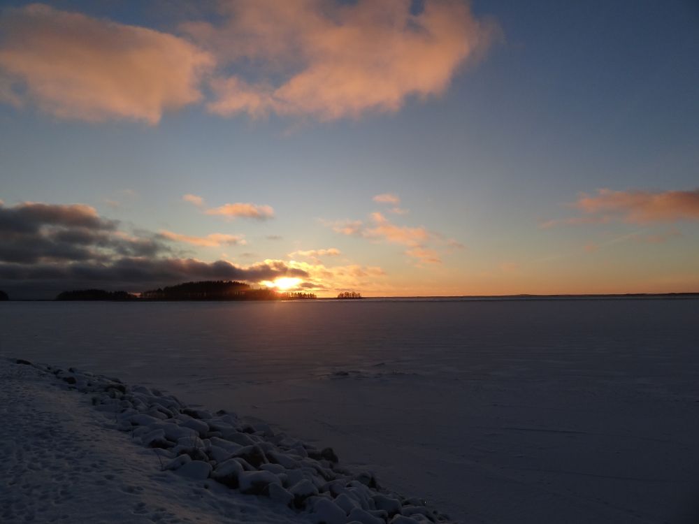 Sun setting behind trees of an island. The island is surrounded by a frozen river with snow on it.