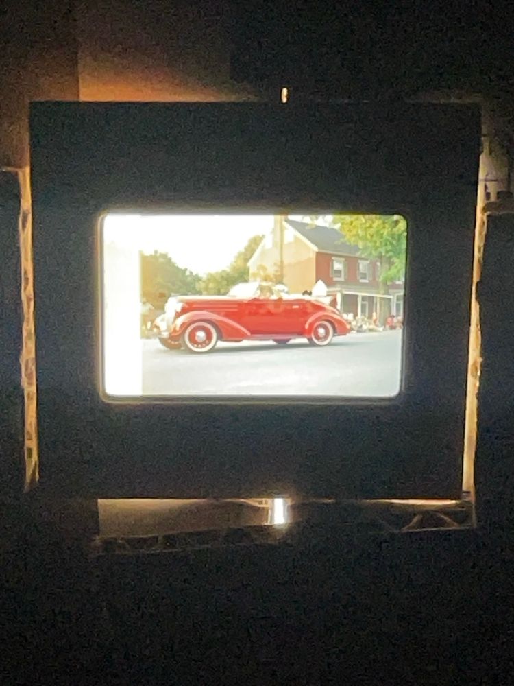 A slide showing a red car from the 1940s being driven in a parade with a house in the background.