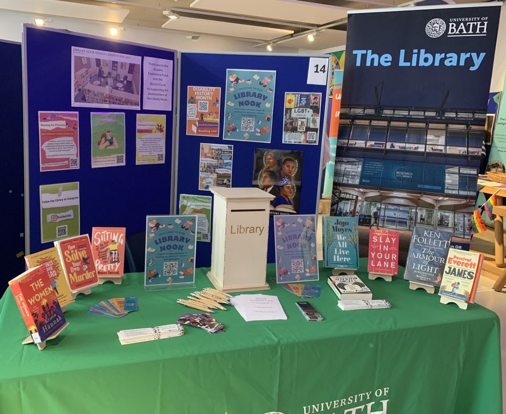 A picture of the Library's stall at the University of Bath Inclusion and Community event. Shows a table covered by a tablecloth with a selection of books on it and a suggestion box. Boards behind the table have posters about the Library Nook and wellbeing reading lists.
