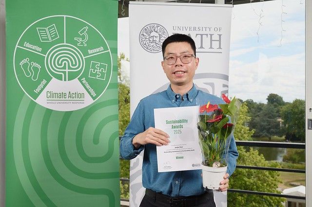 Junjie Shen holding a certificate and plant standing infront of a Climate Action banner and University of Bath banner. Behind is a view across the lake on the University of Bath campus. 