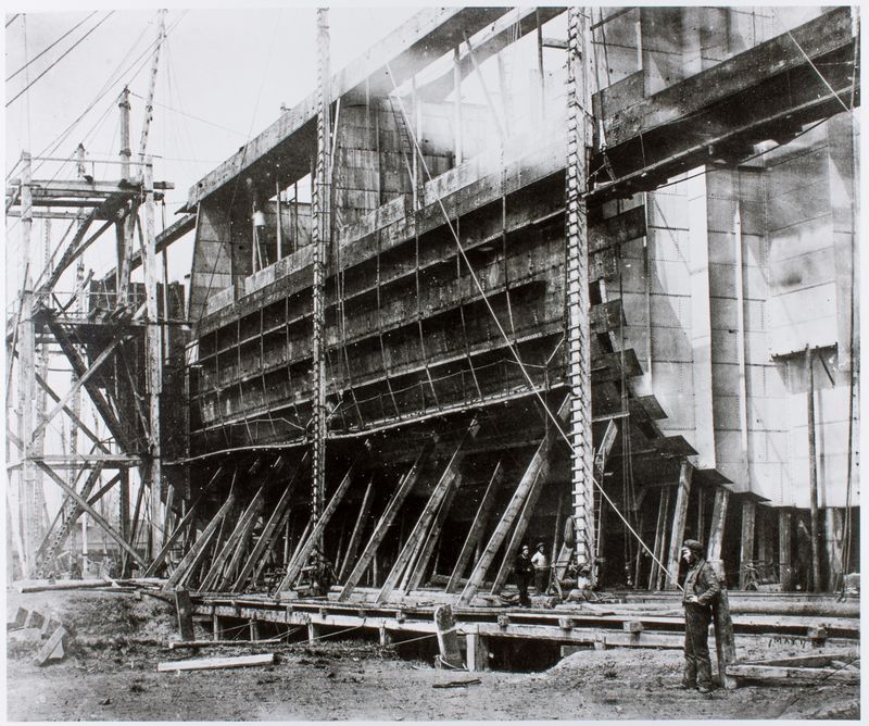 Black and white photograph of the SS Great Eastern being constructed. Section of the ship covered in scaffolding. Man in Victorian dress in the foreground.