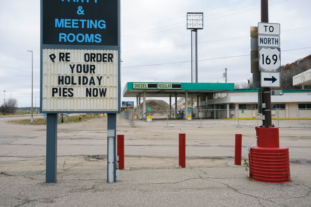 3:2 aspect ratio crop. A sign advertising pre-ordering holiday pies sits beside a highway sign near a highway. Red circular road barriers sit between the signs. A closed gas station with green trim sits behind these two signs.