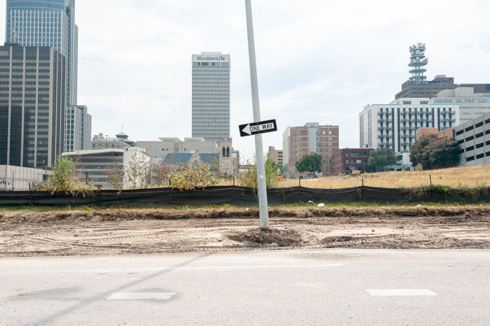 A one way street sign sits at a slant in the center of the image amidst a construction area. A vacant lot and the Omaha skyline lie behind. 