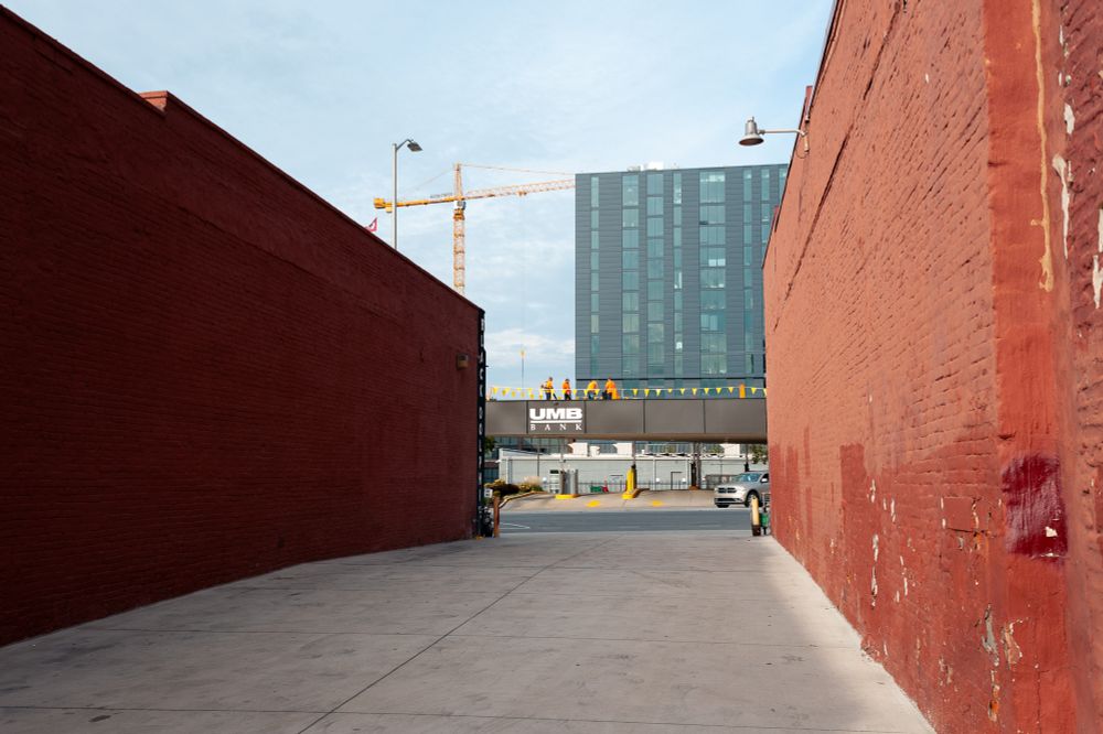 Four workers in orange on a raised platform above the entrance to a bank, viewed through the frame of the orange brick walls of an alley.