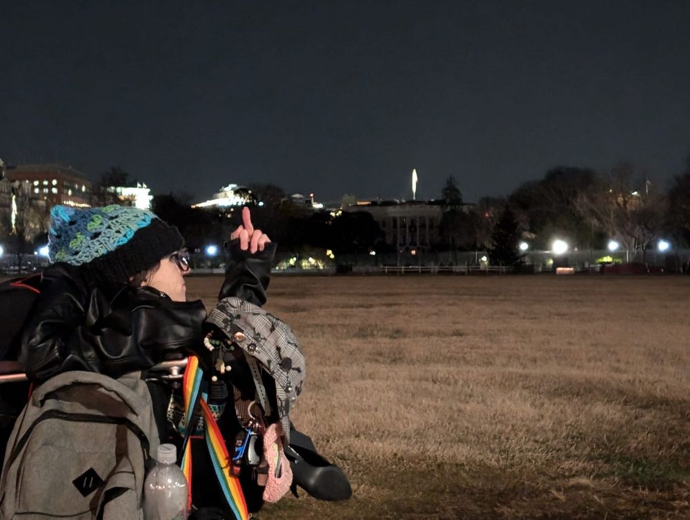Person in a small wheelchair "giving the bird" to a dark white house on a cloudy night.  A rainbow bag strap is clearly visible hanging on the chair.  They are also wearing stilettos, a leather jacket, and a blue crocheted hat with the cat stitch.