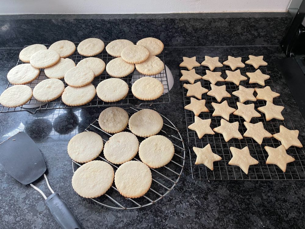 Three cooling racks sit on a black kitchen countertop. Two racks hold round cookies and one holds star-shaped cookies. They are fresh out of the oven, and you can almost smell them through the screen. Nobody, and I mean NOBODY ever guesses that they are gluten-free because they are so damned delicious. (The secret ingredient is cream cheese. Who knew?)
