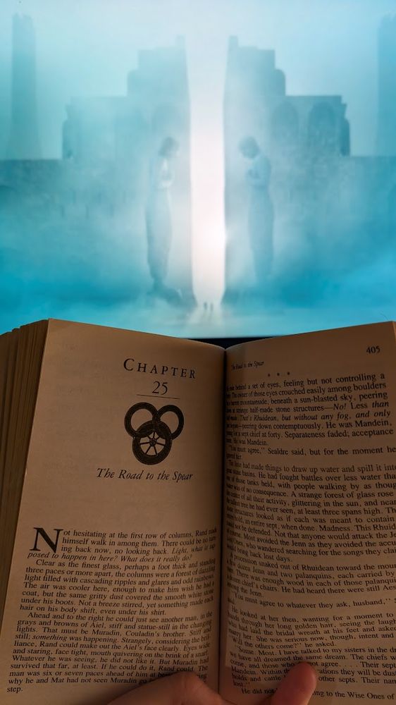 A photo of an open book, The Shadow Rising chapter 25, in front of a still frame from episode 4 of The Wheel of Time season 3. The frame shows a massive gate framed by statues, with two tiny silhouetted figures entering.
